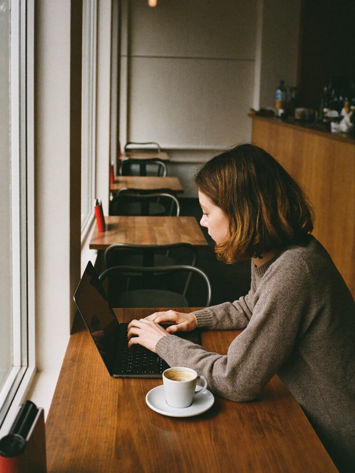 Person sitzt am Fenstertisch eines Cafés mit Laptop und Kaffeetasse, weiches Morgenlicht
